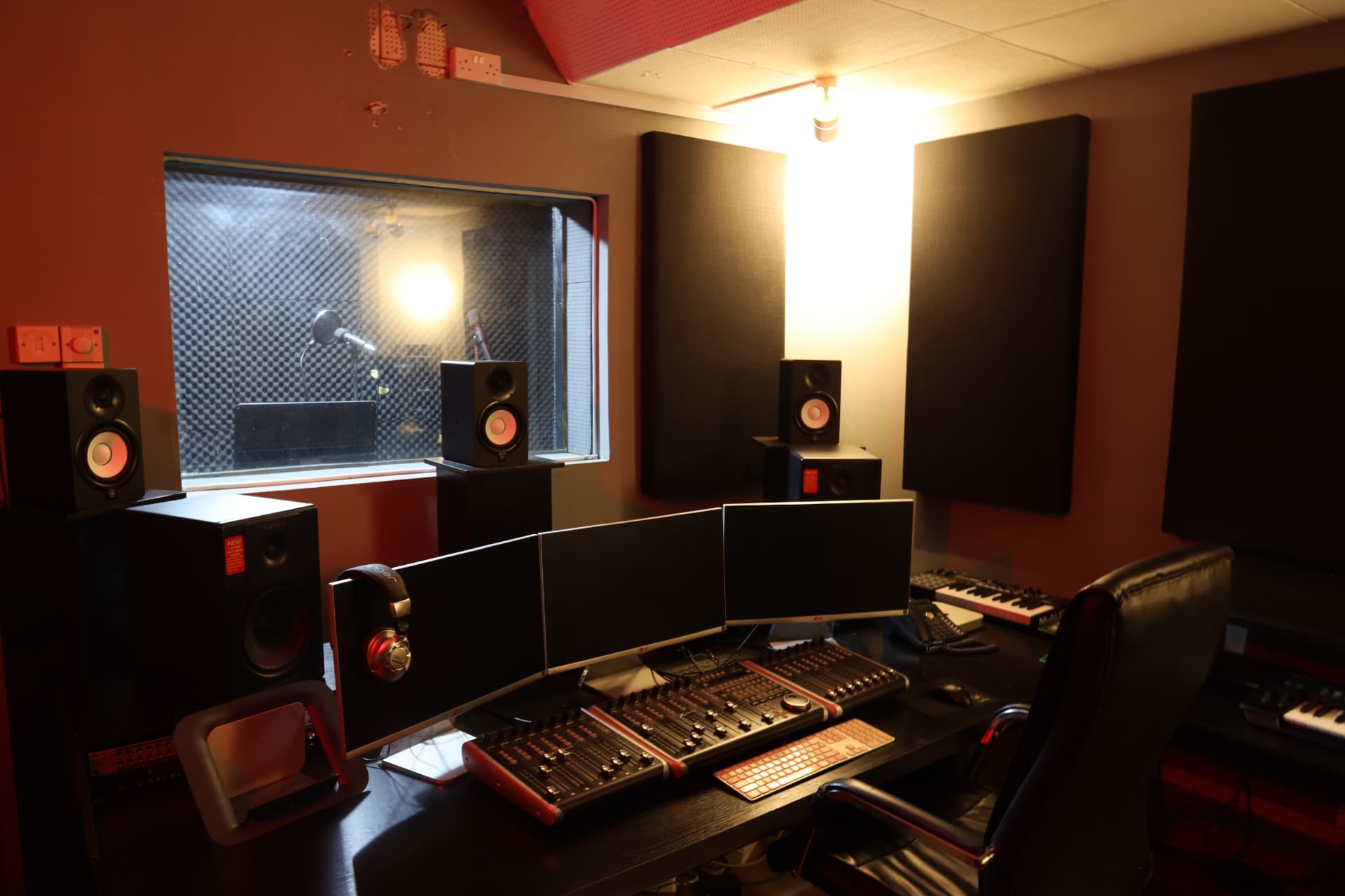 Sound Suite 2 control room with triple monitors, multi-fader mixing console and window into vocal booth