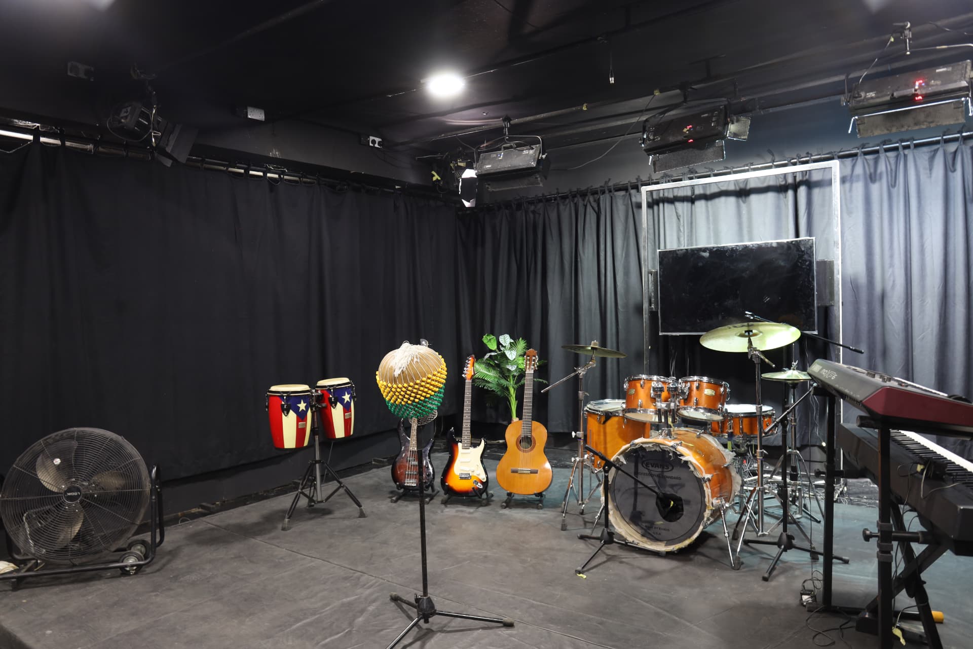 Studio floor stage framed by black curtains, with congas, guitars, bass, drum kit, keyboards and a TV monitor